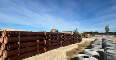 Stacks of vitrified clay drainage pipes and concrete fittings at a construction site under clear skies.