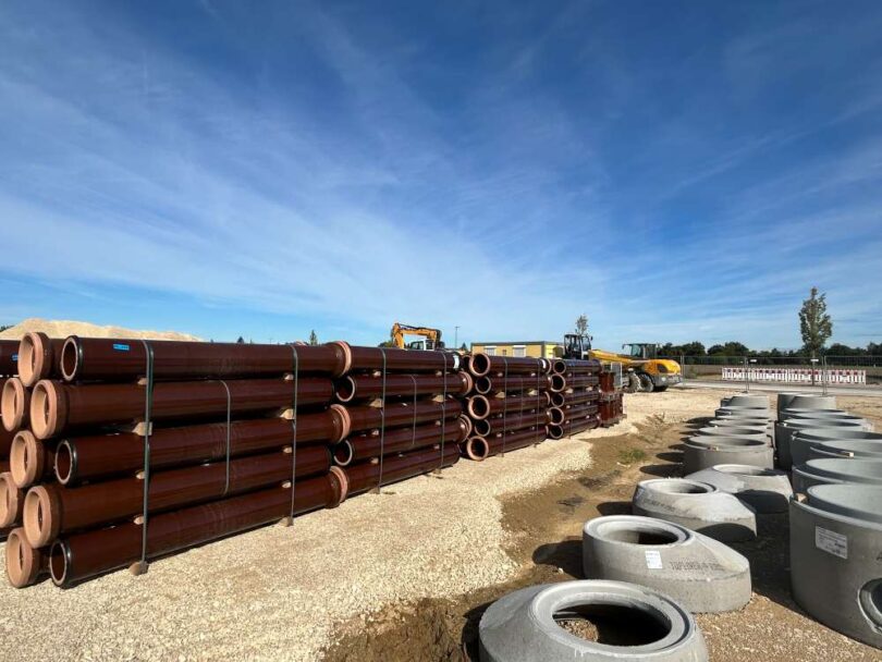 Stacks of vitrified clay drainage pipes and concrete fittings at a construction site under clear skies.