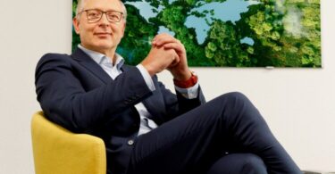 Jürgen von Hollen seated in an office chair with a forest-and-lake artwork behind him.