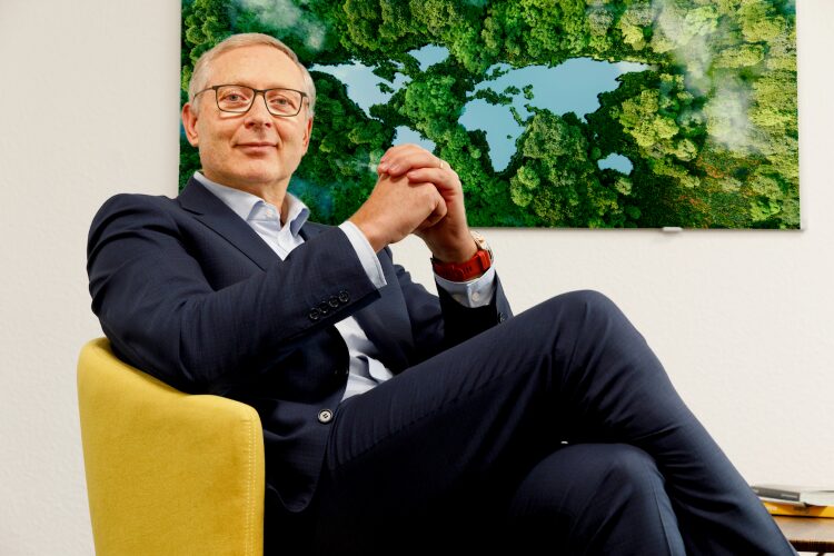 J&uuml;rgen von Hollen seated in an office chair with a forest-and-lake artwork behind him.
