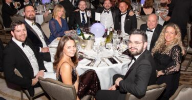 Group of people dressed in formal evening wear seated around a table at an awards ceremony.