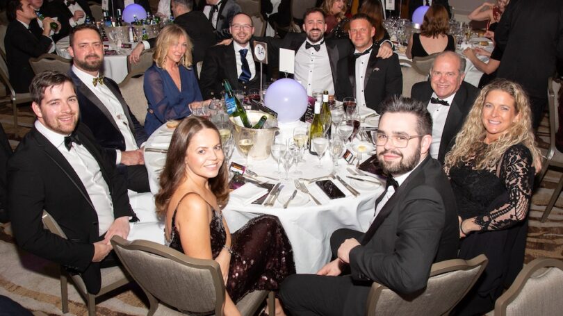Group of people dressed in formal evening wear seated around a table at an awards ceremony.