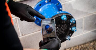 Worker using a smartphone to photograph a blue pipe flange during pipeline quality assurance