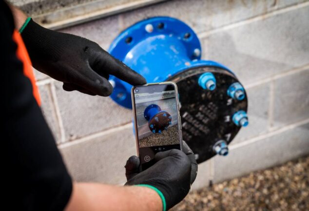 Worker using a smartphone to photograph a blue pipe flange during pipeline quality assurance