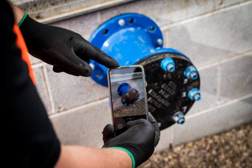 Worker using a smartphone to photograph a blue pipe flange during pipeline quality assurance