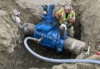 Worker installing a blue insertion valve on a large-diameter fire suppression pipeline in an excavated trench