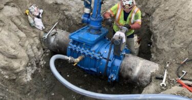 Worker installing a blue insertion valve on a large-diameter fire suppression pipeline in an excavated trench