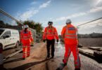 Three water industry workers wearing orange high-visibility clothing and hard hats walking along a construction site