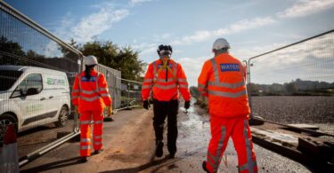 Three water industry workers wearing orange high-visibility clothing and hard hats walking along a construction site