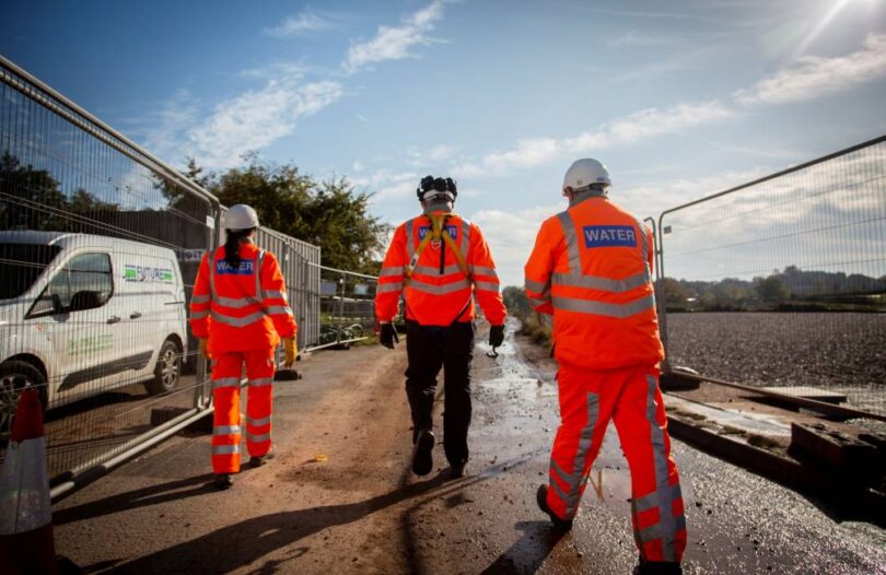 Three water industry workers wearing orange high-visibility clothing and hard hats walking along a construction site