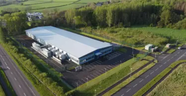 Aerial view of a large industrial building with a pale roof, rooftop-style cooling units, car park and perimeter fence beside