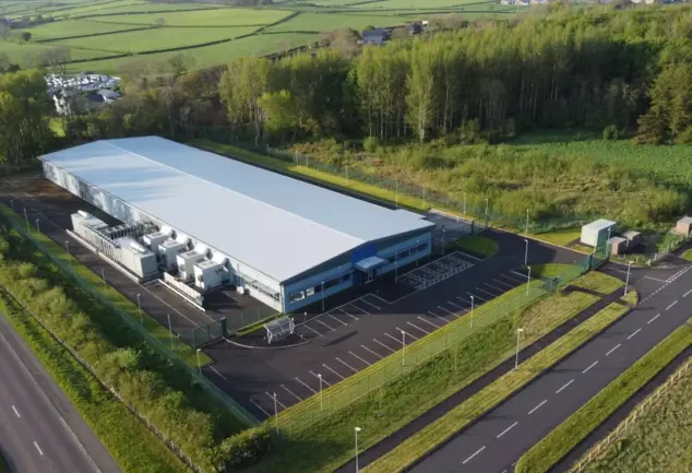 Aerial view of a large industrial building with a pale roof, rooftop-style cooling units, car park and perimeter fence beside