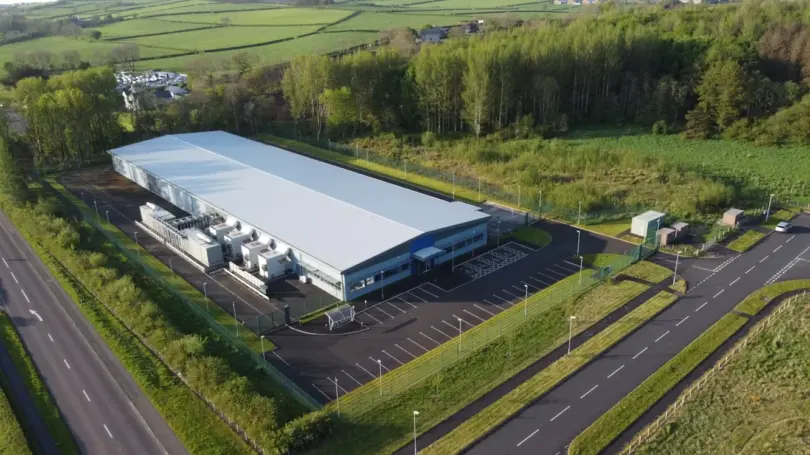 Aerial view of a large industrial building with a pale roof, rooftop-style cooling units, car park and perimeter fence beside