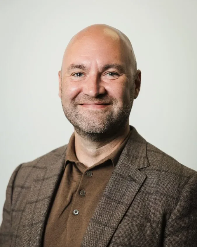 Smiling bald man with a graying stubble beard wearing a brown checked blazer over a brown polo shirt against a plain light ba