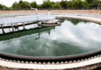 Circular clarifier tank filled with water at a modern urban wastewater treatment plant, with trees behind