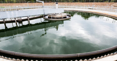 Circular clarifier tank filled with water at a modern urban wastewater treatment plant, with trees behind