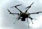Multirotor drone with six arms and a dark sensor payload hovering against a cloudy gray sky