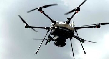 Multirotor drone with six arms and a dark sensor payload hovering against a cloudy gray sky