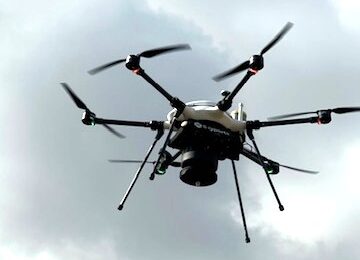 Multirotor drone with six arms and a dark sensor payload hovering against a cloudy gray sky