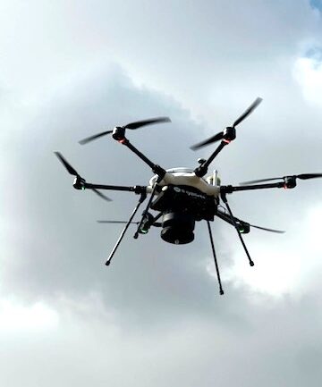 Multirotor drone with six arms and a dark sensor payload hovering against a cloudy gray sky