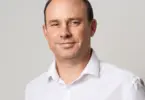 Smiling middle-aged man with short dark hair and a receding hairline, wearing a white collared shirt against a plain light gr