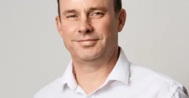 Smiling middle-aged man with short dark hair and a receding hairline, wearing a white collared shirt against a plain light gr
