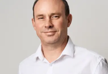 Smiling middle-aged man with short dark hair and a receding hairline, wearing a white collared shirt against a plain light gr