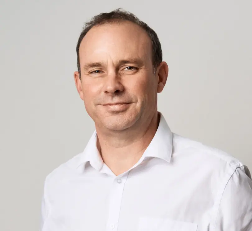 Smiling middle-aged man with short dark hair and a receding hairline, wearing a white collared shirt against a plain light gr