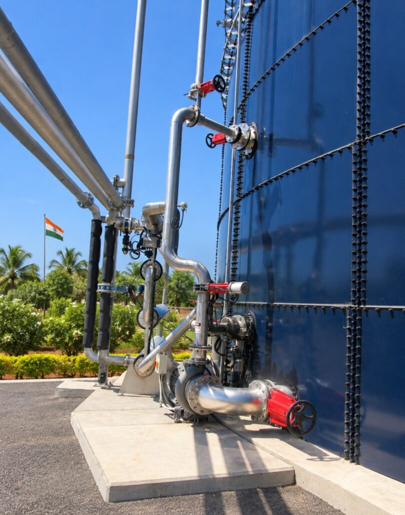 Stainless steel pipes, pump and red-handled valves along a large blue glass-fused steel tank, with an Indian flag in the back