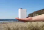 Open palm holding a small white handheld device with embossed lettering, with a beach, sea and grassy headland behind