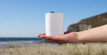 Open palm holding a small white handheld device with embossed lettering, with a beach, sea and grassy headland behind