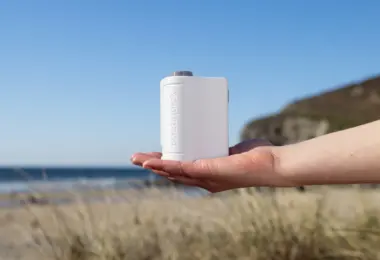Open palm holding a small white handheld device with embossed lettering, with a beach, sea and grassy headland behind