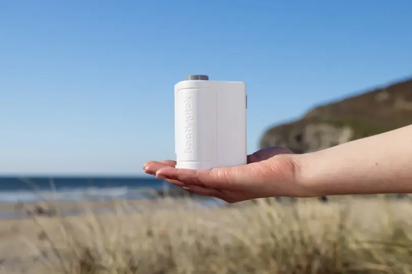 Open palm holding a small white handheld device with embossed lettering, with a beach, sea and grassy headland behind