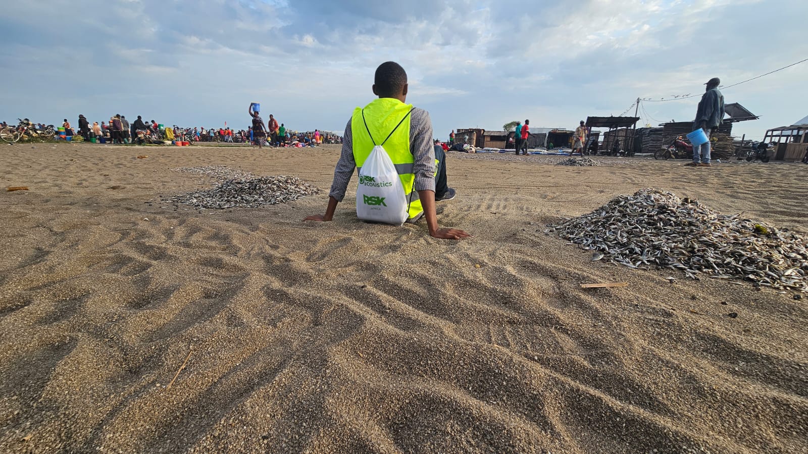 Person in a hi-vis vest and RSK Acoustics backpack sits on sand facing a busy lakeshore fish market with small fish drying in