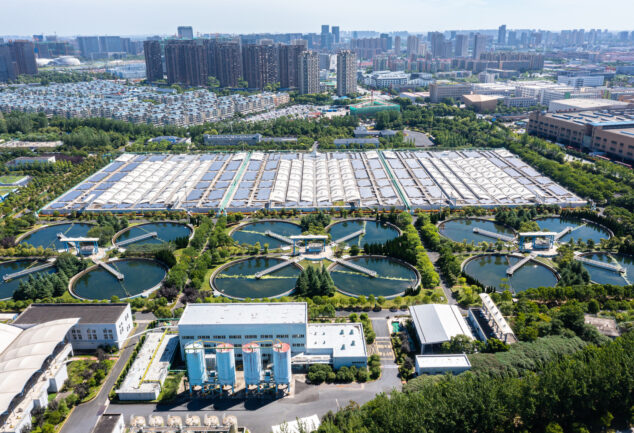 Sewage treatment plant with a solar power station, showing water treatment tanks and rows of solar panels.