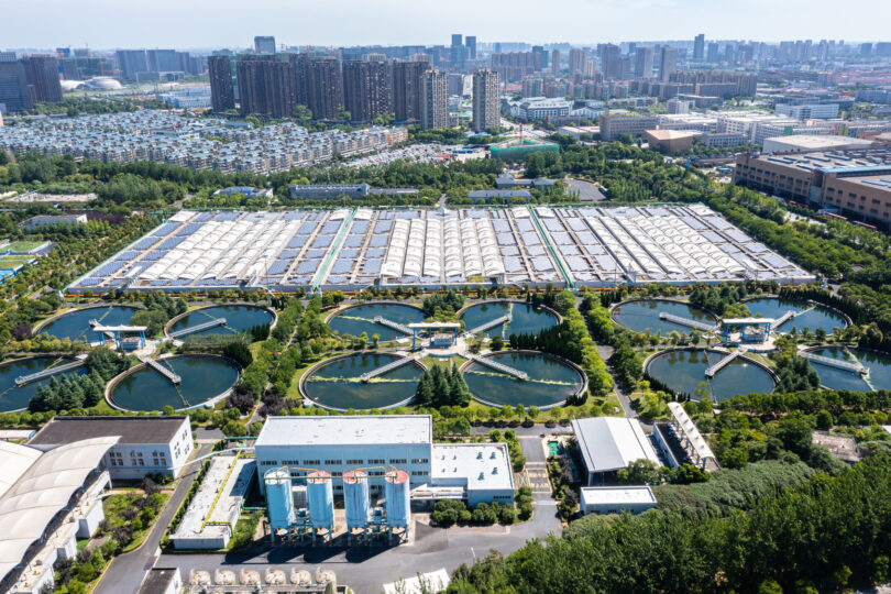 Sewage treatment plant with a solar power station, showing water treatment tanks and rows of solar panels.