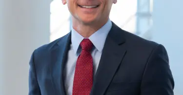 Smiling man with short brown hair in a navy suit, light blue shirt and red patterned tie, standing before a bright window