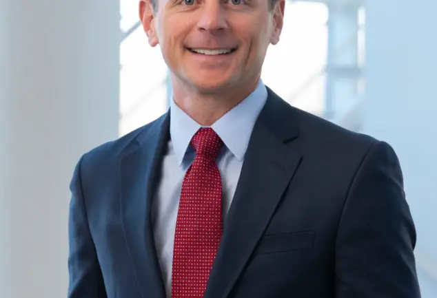 Smiling man with short brown hair in a navy suit, light blue shirt and red patterned tie, standing before a bright window