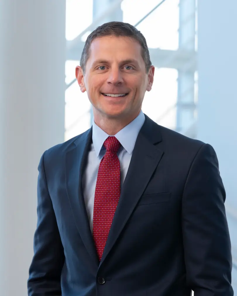 Smiling man with short brown hair in a navy suit, light blue shirt and red patterned tie, standing before a bright window