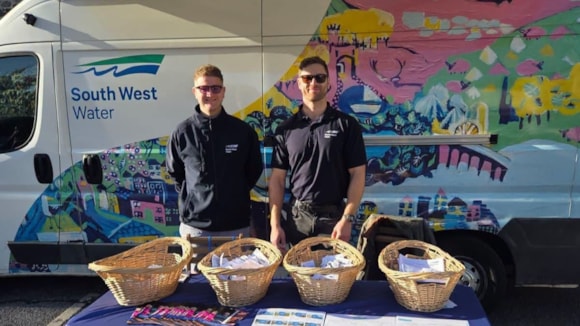 Two South West Water representatives standing behind a table with wicker baskets of information packs beside a branded van