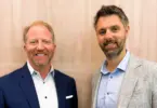 Two smiling men in blazers and collared shirts stand side by side in front of a light wood-paneled wall.