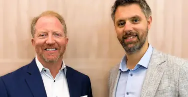 Two smiling men in blazers and collared shirts stand side by side in front of a light wood-paneled wall.