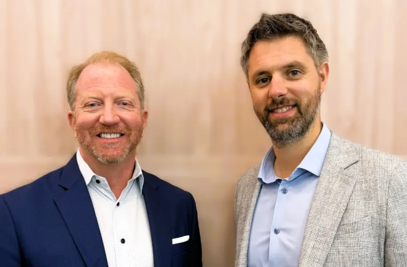 Two smiling men in blazers and collared shirts stand side by side in front of a light wood-paneled wall.