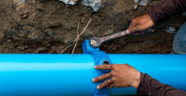 A man uses a large wrench on water pipes, with metal pipework visible around him.