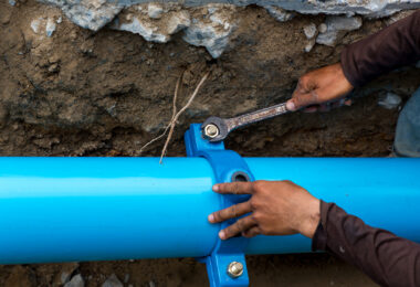 A man uses a large wrench on water pipes, with metal pipework visible around him.