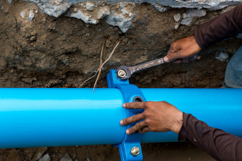 A man uses a large wrench on water pipes, with metal pipework visible around him.