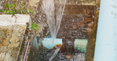 A broken pipe lying in a hole in a roadside ditch, with water flowing out of it.