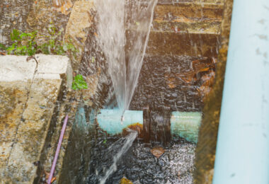 A broken pipe lying in a hole in a roadside ditch, with water flowing out of it.
