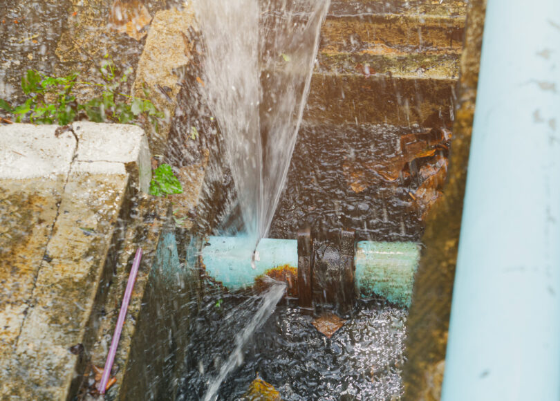 A broken pipe lying in a hole in a roadside ditch, with water flowing out of it.