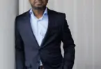 Man in a navy blazer and light blue open-collar shirt leans against a wall, one hand in his pocket, smiling slightly at the c